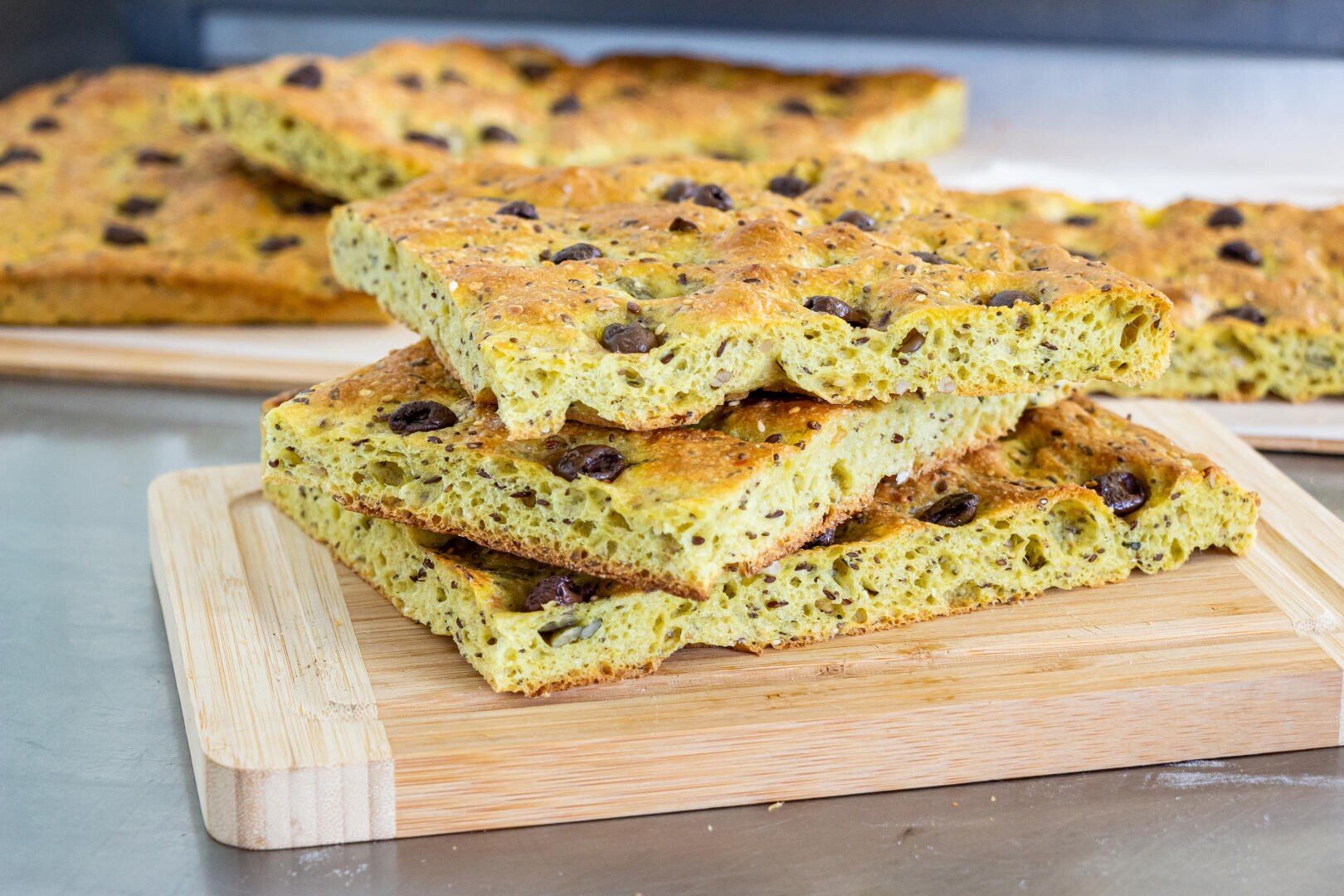 Three rectangular slices of focaccia bread with olives are stacked on a wooden cutting board, with more pieces of focaccia visible in the background on a metal surface.