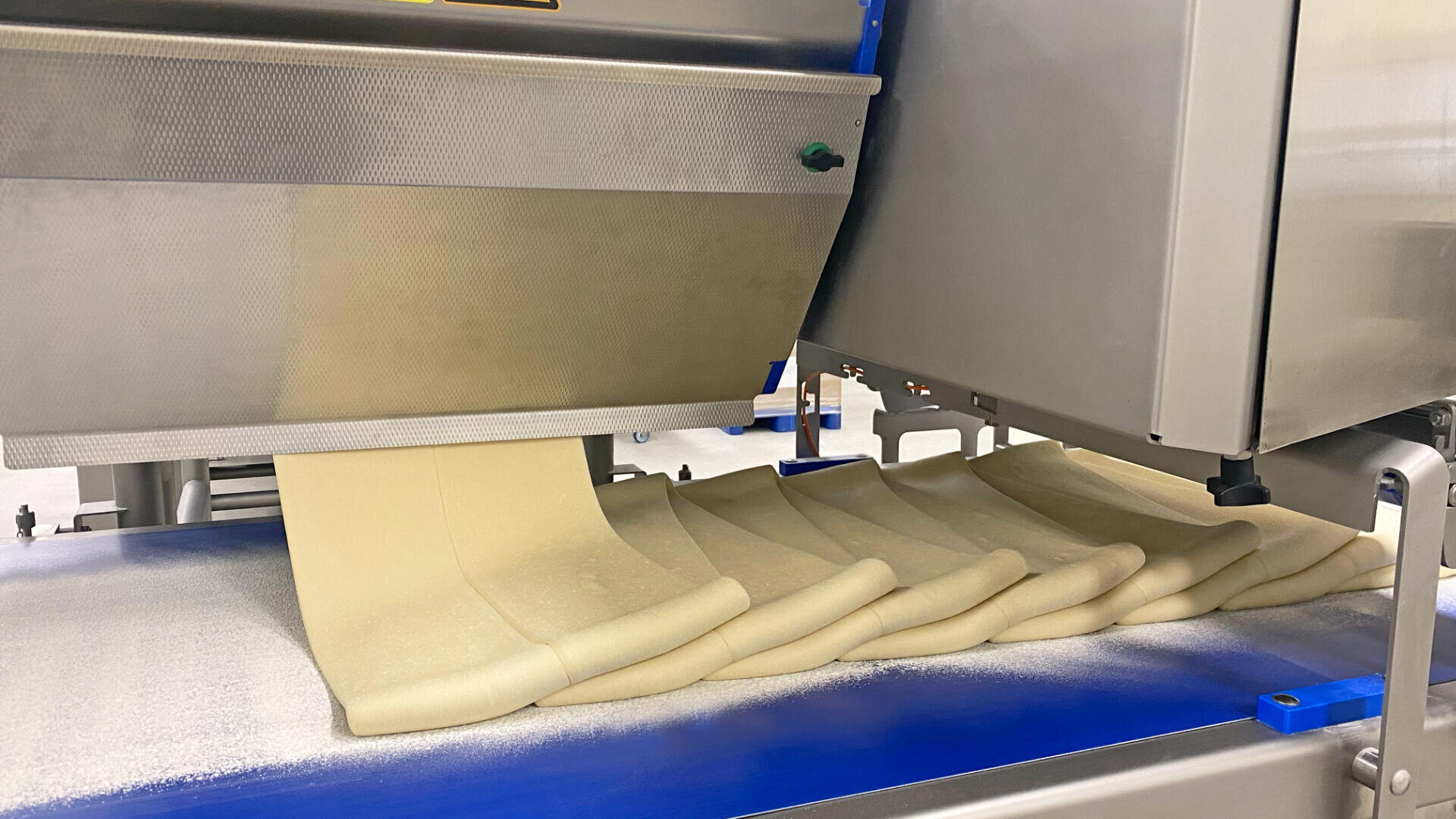 Sheets of dough being fed out of a large industrial machine onto a conveyor belt, with some flour dusted on the surface.