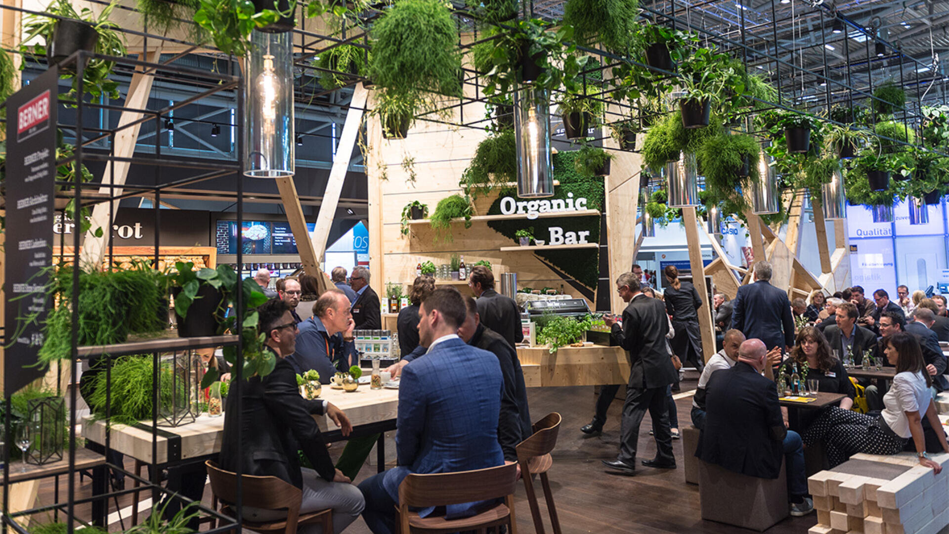 Ein belebtes Innencafé mit vielen Menschen, die an Tischen sitzen und stehen. Der Raum ist mit hängenden Grünpflanzen dekoriert und trägt ein Schild mit der Aufschrift Organic Bar. Die Atmosphäre ist lebendig und modern.