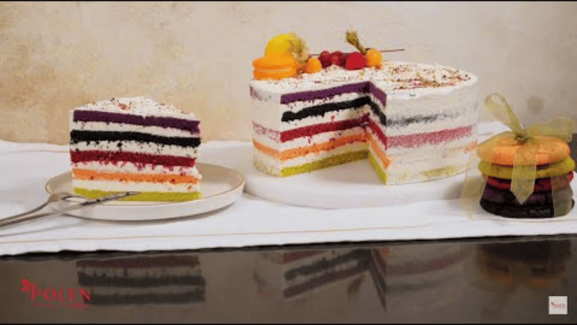 Auf einem weißen Tuch liegt ein bunter, mehrschichtiger Kuchen, von dem ein Stück auf einem Teller serviert wird. Daneben liegt ein Stapel bunter Kekse, die mit einer Schleife zusammengebunden sind. Die Torte ist mit Früchten dekoriert.