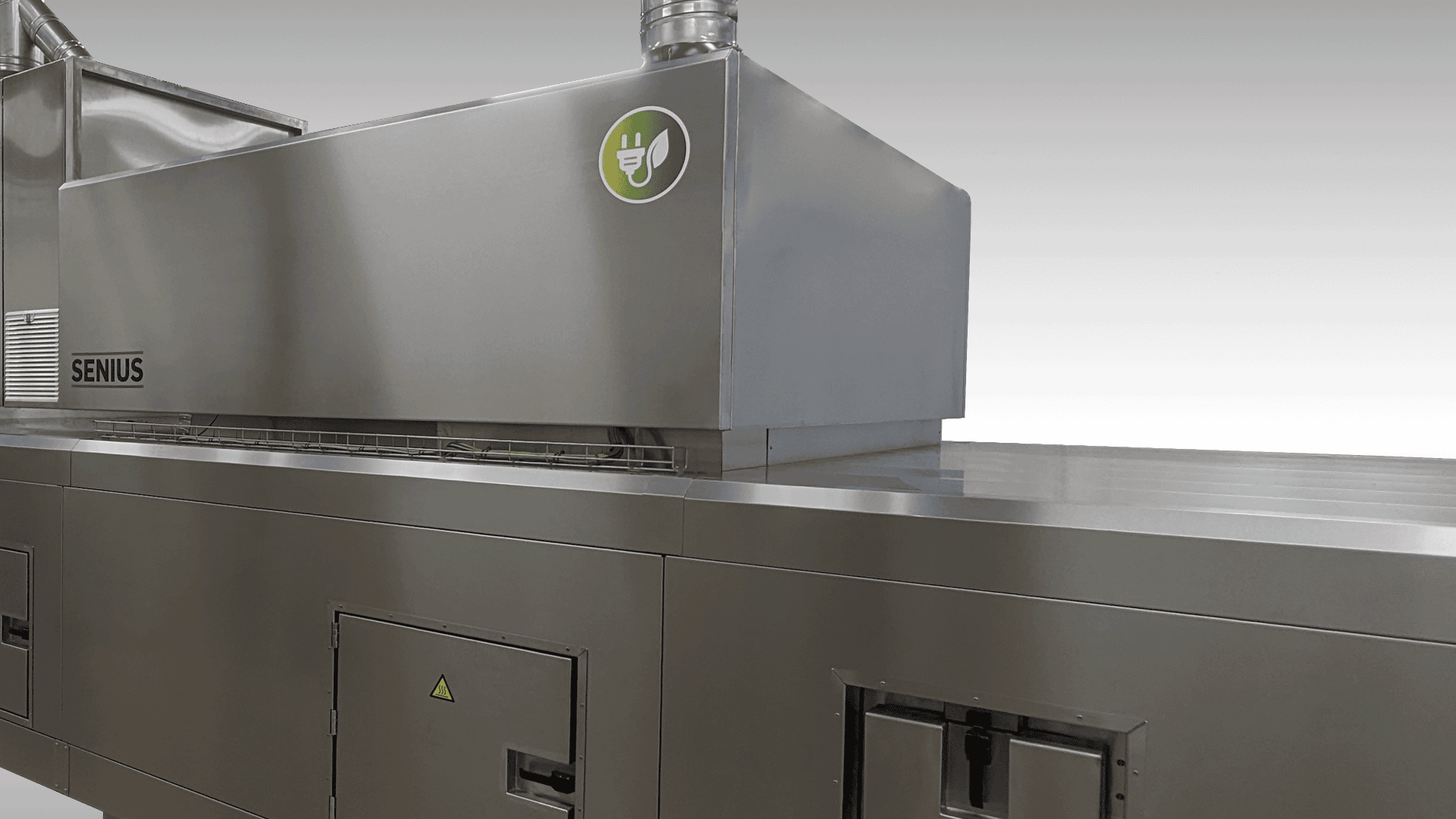 A large stainless steel industrial machine with a ventilation duct and an energy efficiency sticker on its upper section, labeled SENIUS, set against a plain light background.