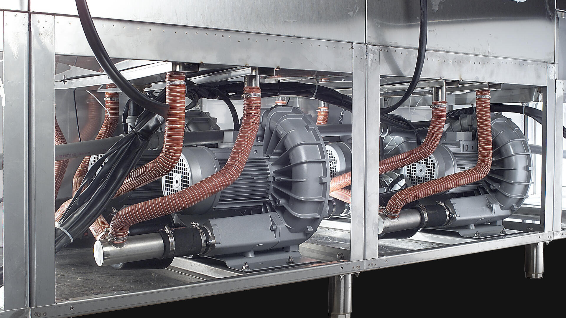 A close-up view of industrial machinery with large electric motors, orange flexible hoses, and metallic components housed in a stainless steel frame.