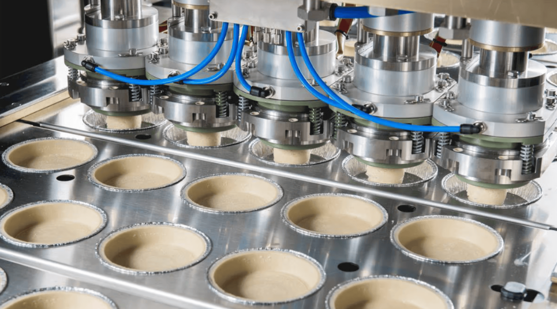 Nahaufnahme einer Maschine zur Herstellung von Lebensmitteln, die Teig in Reihen von runden Metallschalen auf einem Fließband ausgibt. Über den Schalen sind blaue Kabel und präzise Düsen zu sehen.