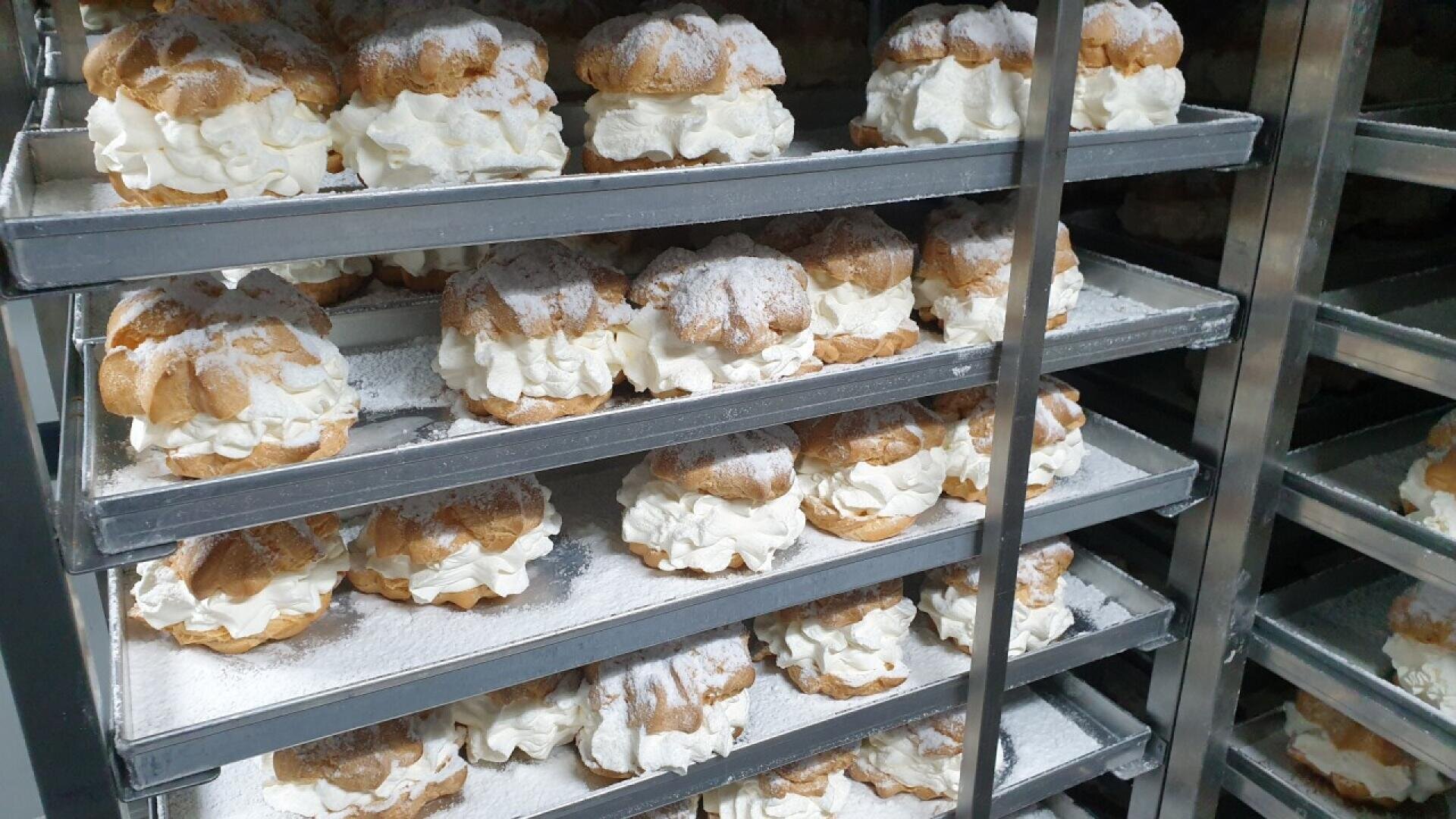 Multiple trays filled with cream puffs topped with powdered sugar are stacked on metal racks, likely in a bakery or kitchen setting.