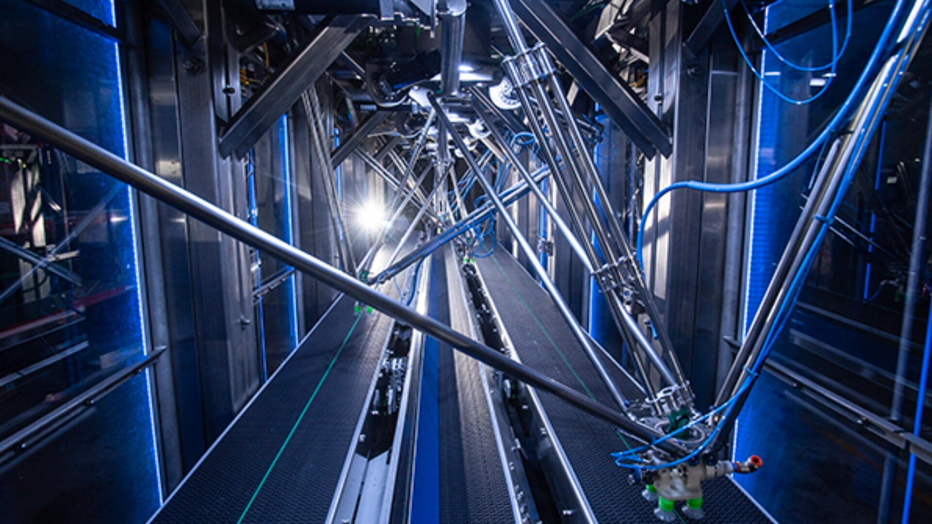 A brightly lit, futuristic industrial corridor filled with robotic arms and cables extends into the distance, reflecting blue light off metallic surfaces. The scene suggests advanced automation or a high-tech manufacturing process.