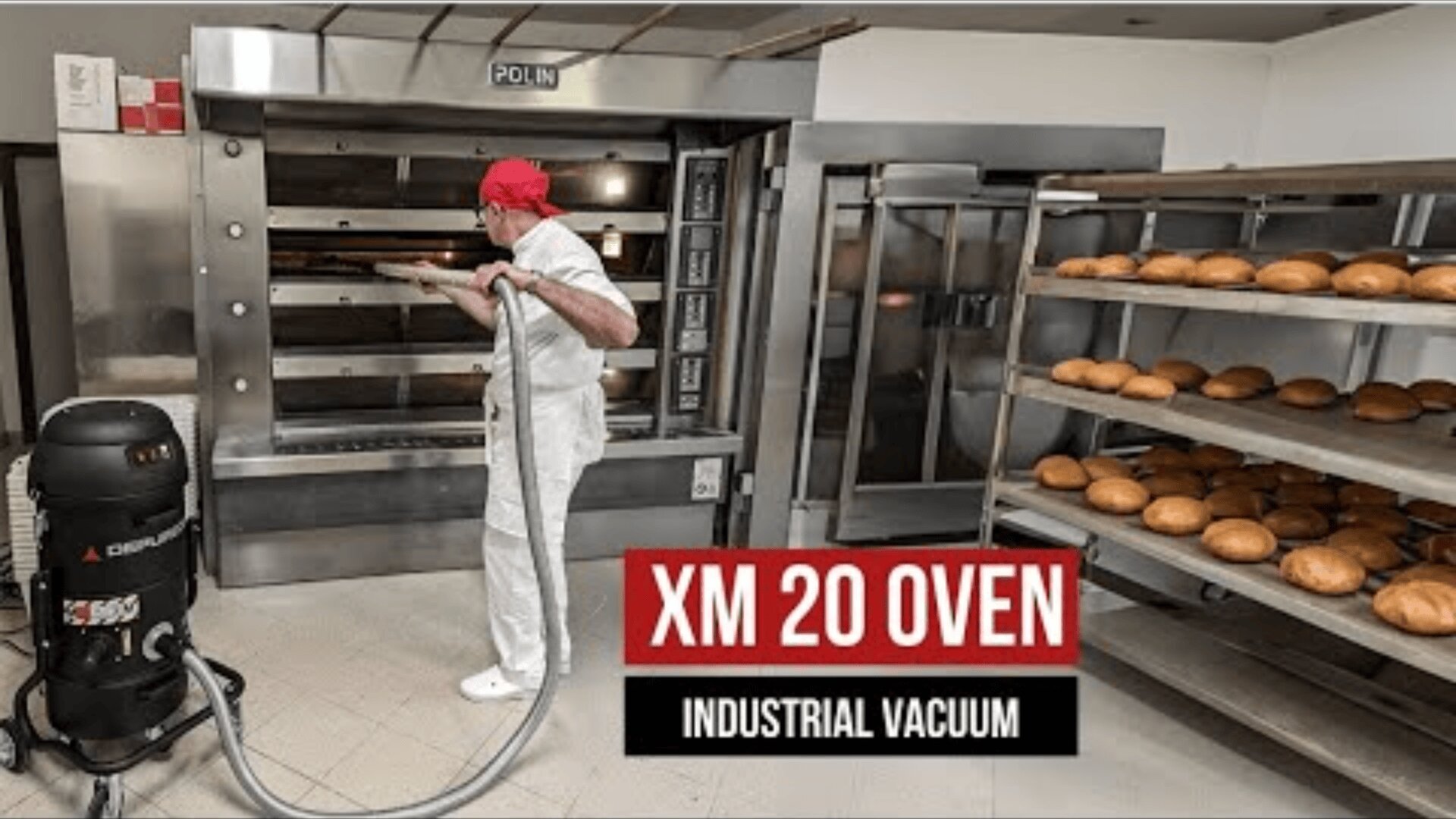 Ein Bäcker in weißer Uniform und roter Mütze reinigt mit einem Staubsauger einen großen Industrieofen, in dessen Nähe sich Regale mit frisch gebackenem Brot befinden. Der Text lautet "XM 20 OVEN INDUSTRIAL VACUUM.".