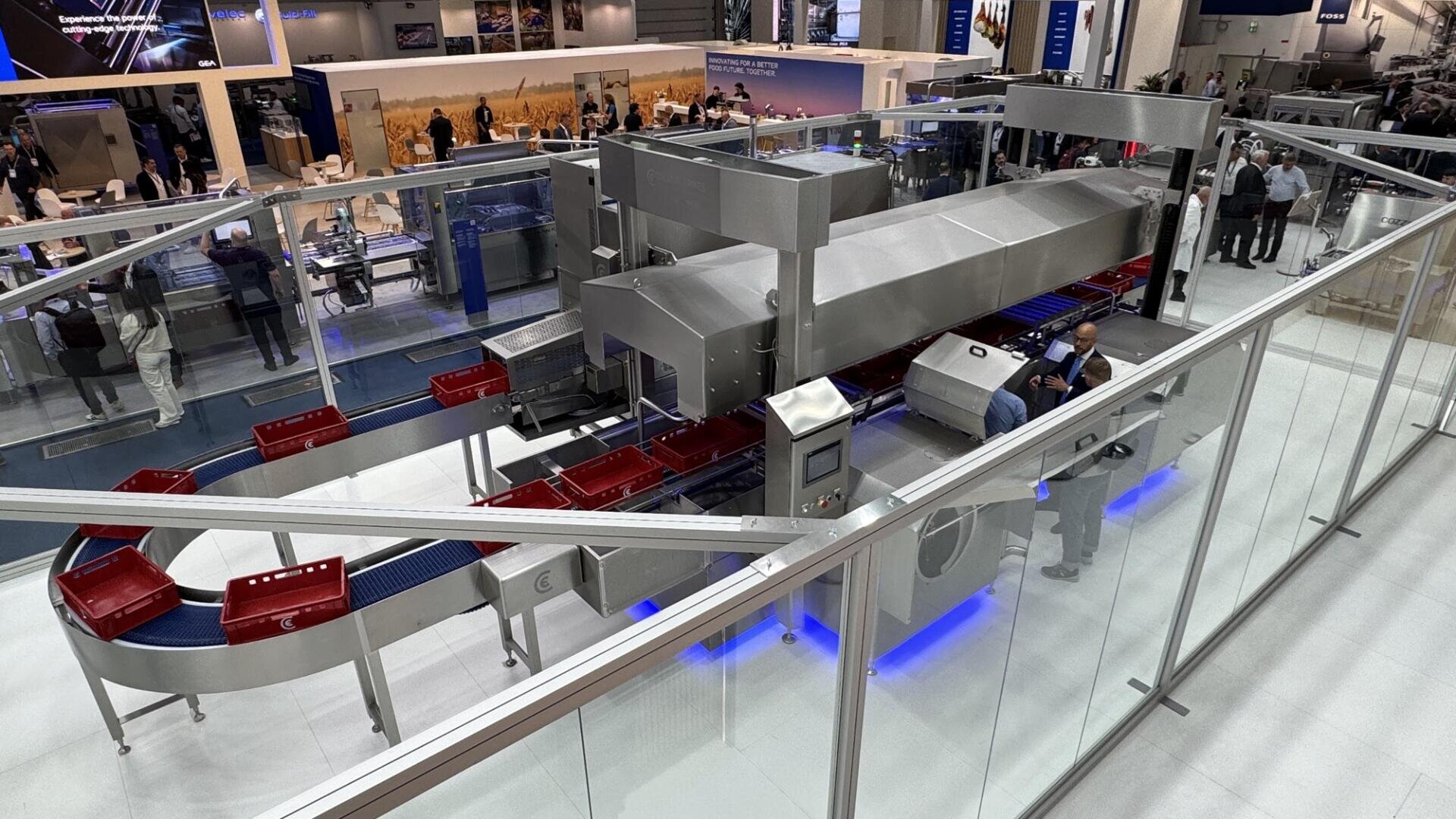 Large industrial food processing machine with a red and silver conveyor belt system on display at a trade show; people observe and interact with the equipment inside a glass-enclosed area.