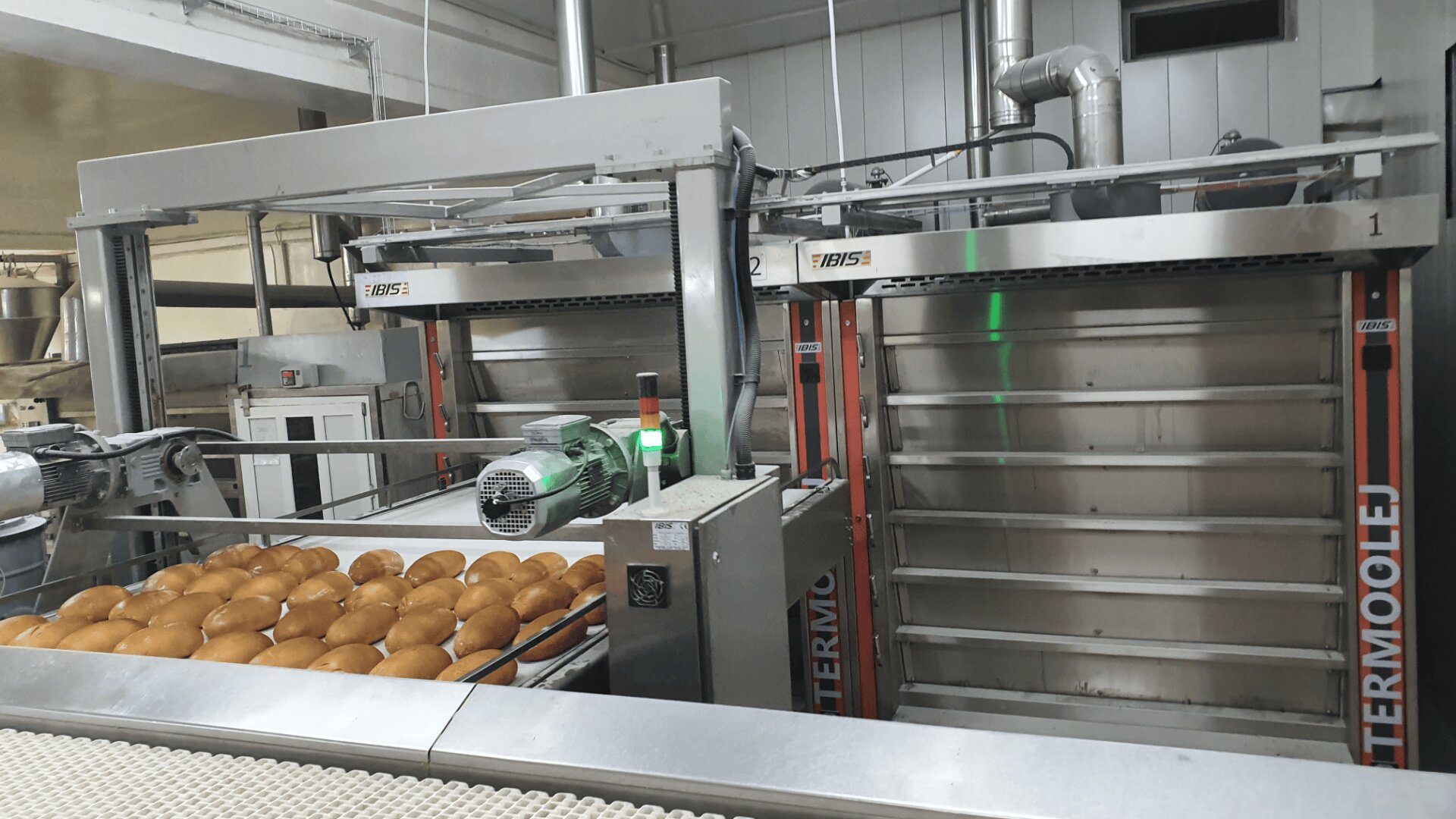 Rows of freshly baked bread rolls are on a conveyor belt inside an industrial bakery. Large metallic ovens and machinery are visible in the background, along with control panels and ventilation pipes.