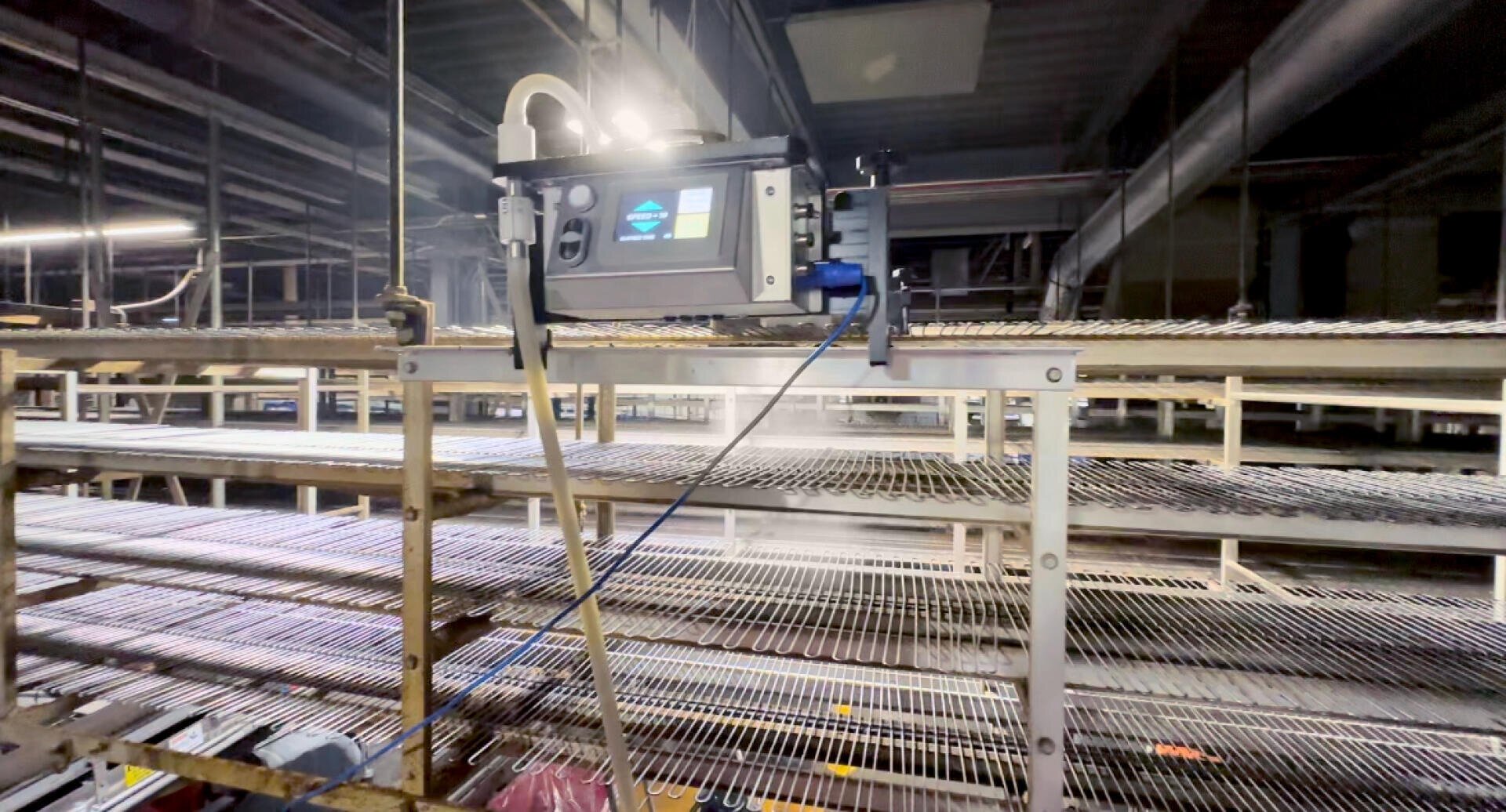 A close-up view of an industrial machine with a screen and cables mounted above empty metal conveyor racks inside a large, dimly lit factory or warehouse.