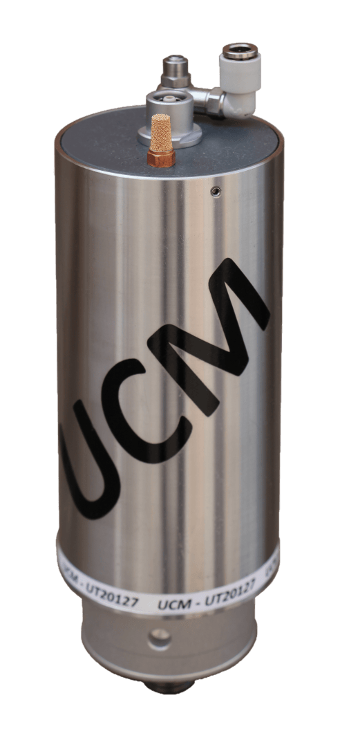 A cylindrical metallic device with the letters UCM printed on the side, featuring connectors and a fitting on the top and a label around the base.