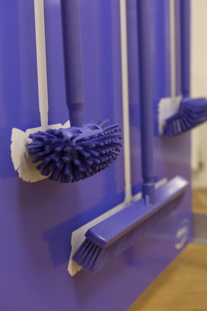 Nahaufnahme von blauen Reinigungsbürsten und Besen, die ordentlich an einem passenden blauen Wandregal mit weißen Haken hängen.