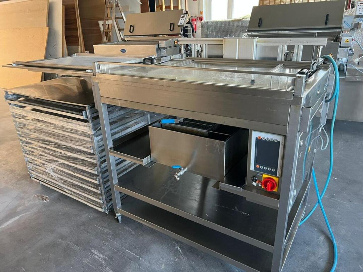 Eine große Industriespüle aus rostfreiem Stahl mit Bedienknöpfen, einem blauen Schlauch und in der Nähe gestapelten Metallregalen, die in einer Werkstatt oder Industrieküche aufgestellt sind.