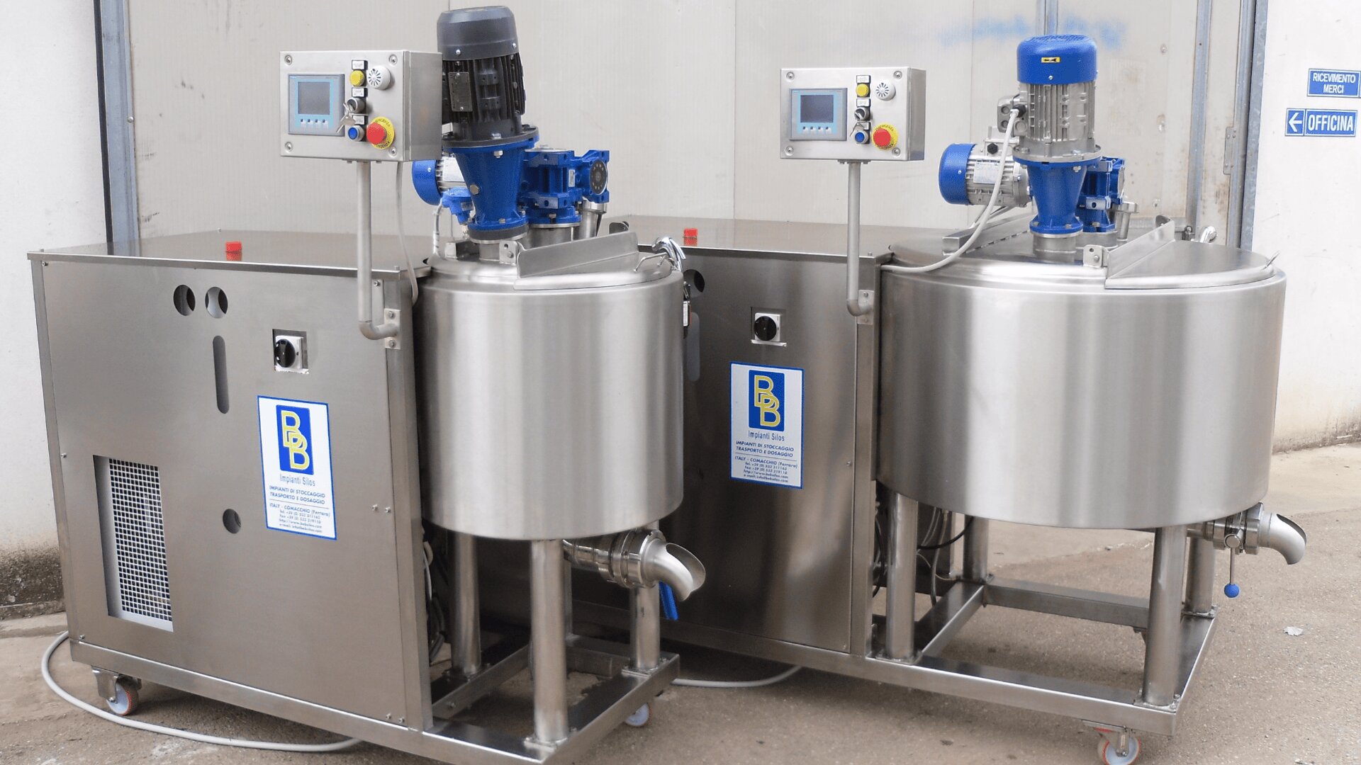 Two large industrial stainless steel mixing machines with control panels, motors, and pipes, placed on wheels in an indoor industrial setting. Each machine has a blue and yellow company logo label on the front.