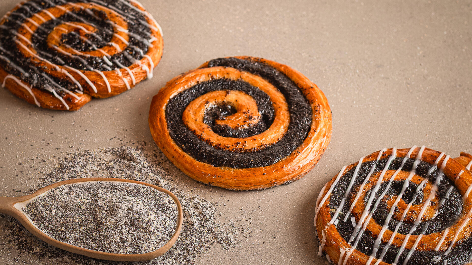 Drei spiralförmige Mohnschnecken mit Zuckerguss liegen auf einem hellbraunen Untergrund. Ein mit gemahlenem Mohn gefüllter Holzlöffel liegt daneben, daneben sind einige Samen verstreut.