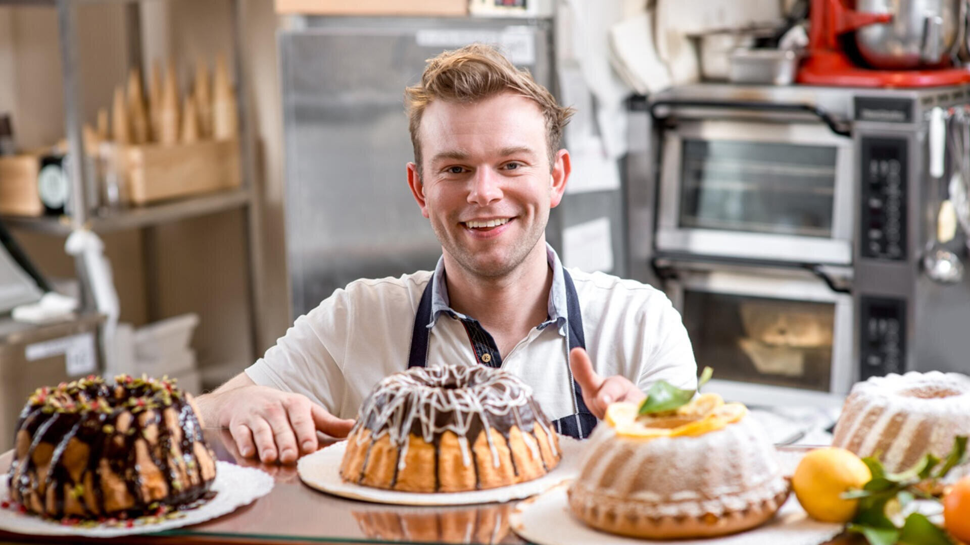 A smiling baker wearing an apron stands behind a counter with three decorated bundt cakes in a bakery kitchen, with ovens and baking tools visible in the background.