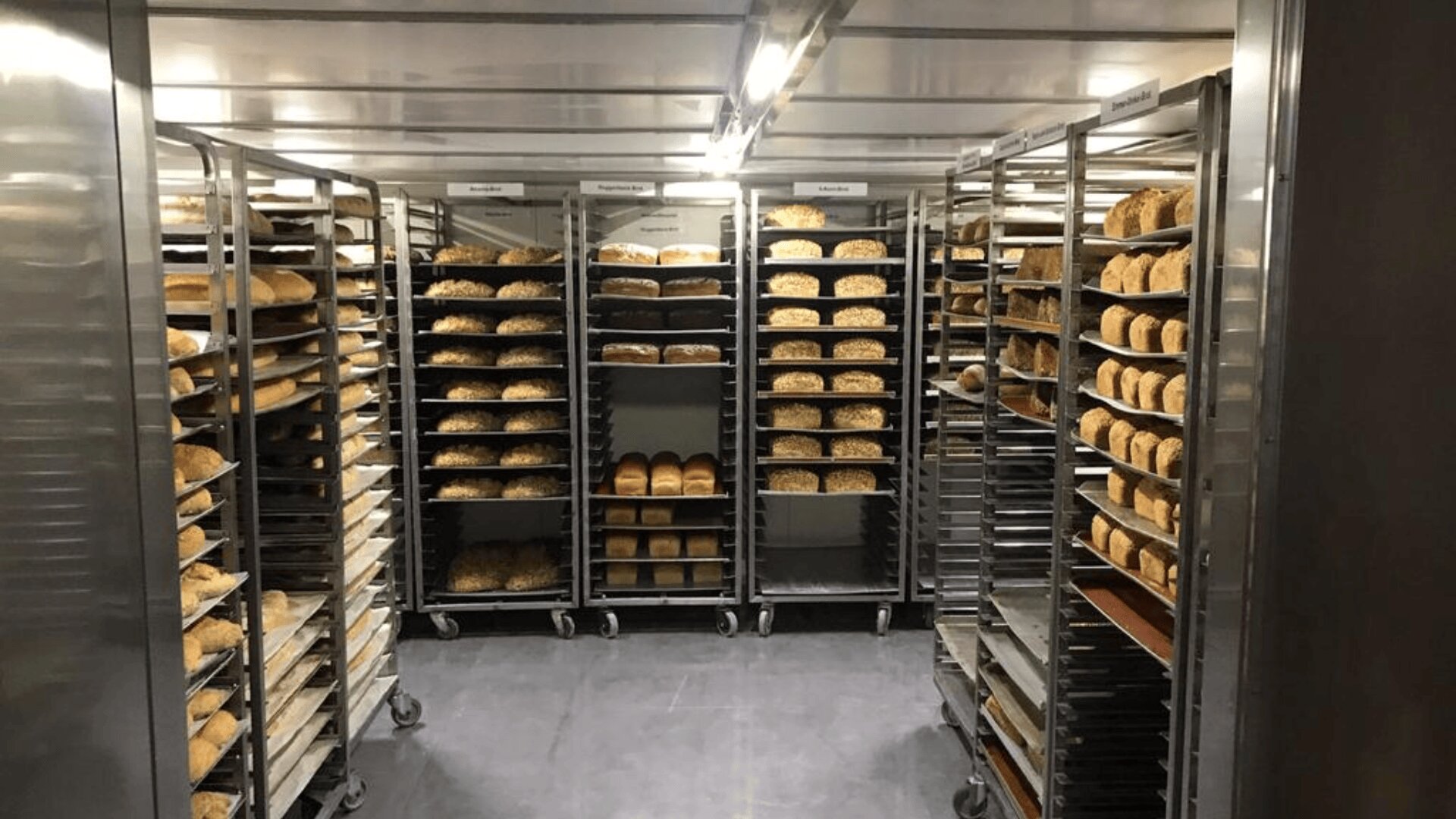 Ein Lagerraum einer gewerblichen Bäckerei mit Metallregalen voller Tabletts mit verschiedenen Brot- und Brötchensorten, die ordentlich angeordnet und in Reihen an den Wänden gestapelt sind.