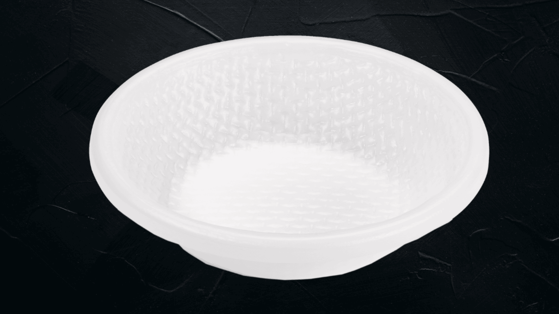 Eine kleine, runde, weiße Kunststoffschale mit einem strukturierten, gewebten Muster auf der Innenseite, die auf einer dunklen, strukturierten Oberfläche steht.
