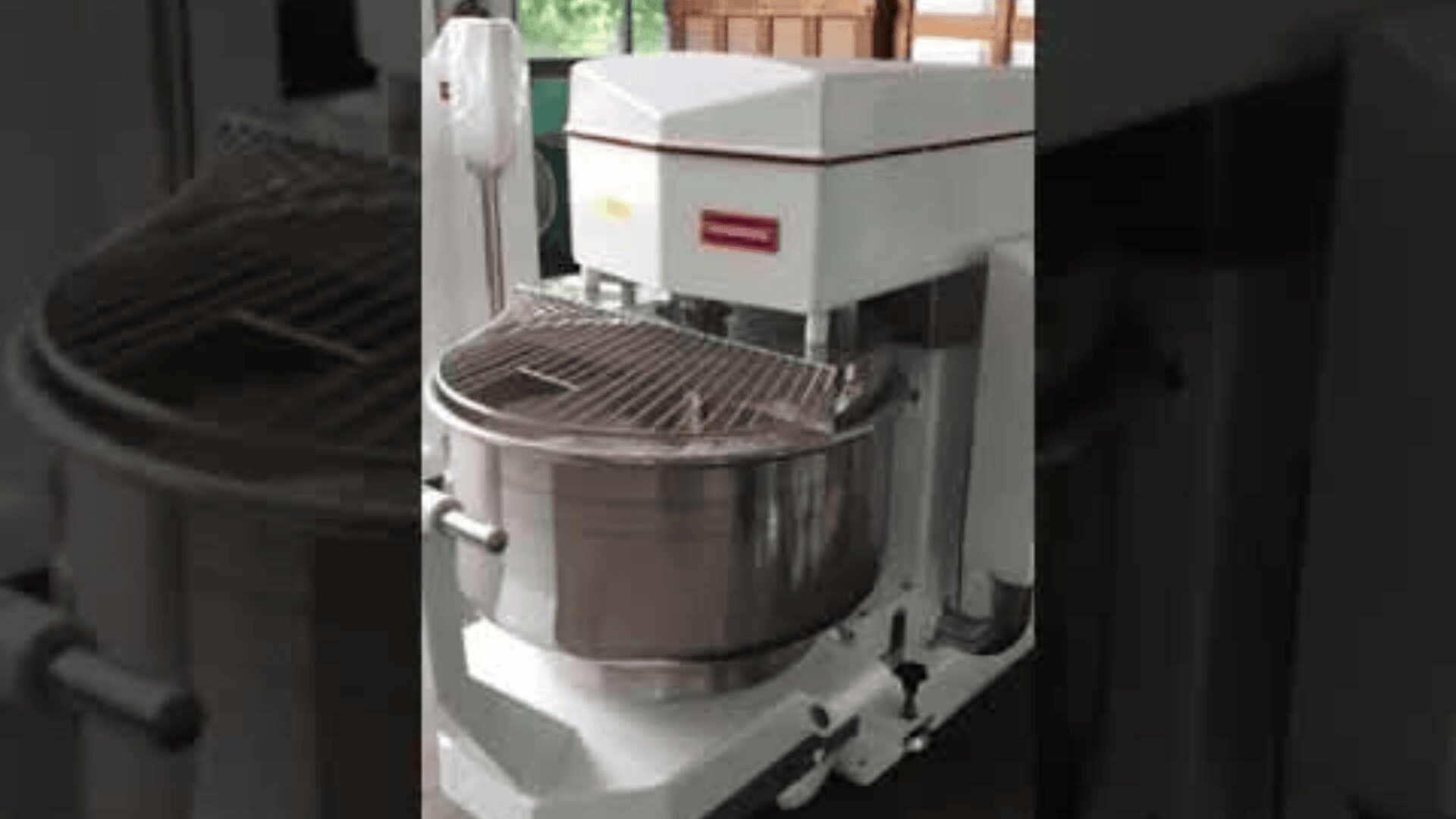 A large industrial dough mixer with a stainless steel bowl and protective metal grate, positioned in a commercial kitchen setting.