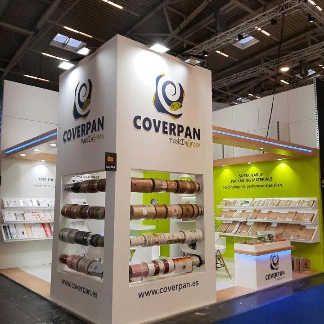 Trade show booth for Coverpan, displaying rolls of packaging material on racks and shelves with samples. The booth features the Coverpan logo, website, and signage about sustainable packaging materials.
