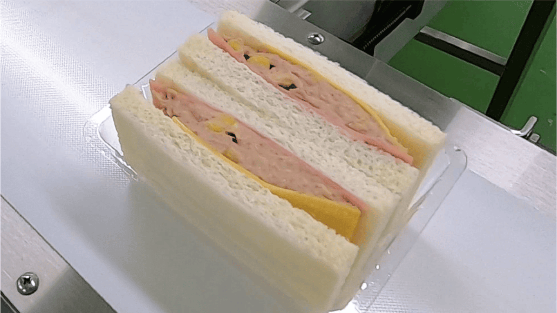 Ein Plastikbehälter mit drei sauber geschnittenen Sandwiches mit Weißbrot, Schinkenscheiben, gelbem Käse und einer Füllung, die Mayonnaise und kleine Gemüsestücke zu enthalten scheint.