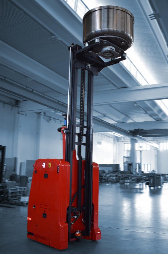 A red industrial forklift lifts a large metal drum high above its head inside a spacious, modern warehouse with bright overhead lighting.