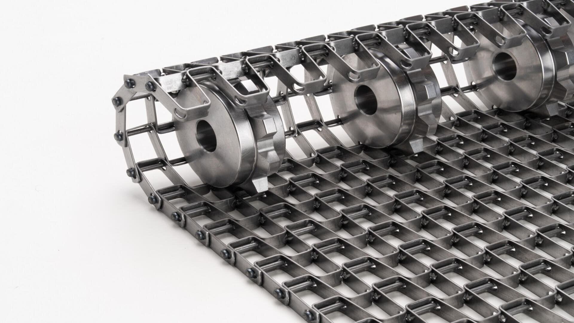 A close-up of a stainless steel conveyor belt with interlocking grid links and cylindrical rollers, photographed against a white background.