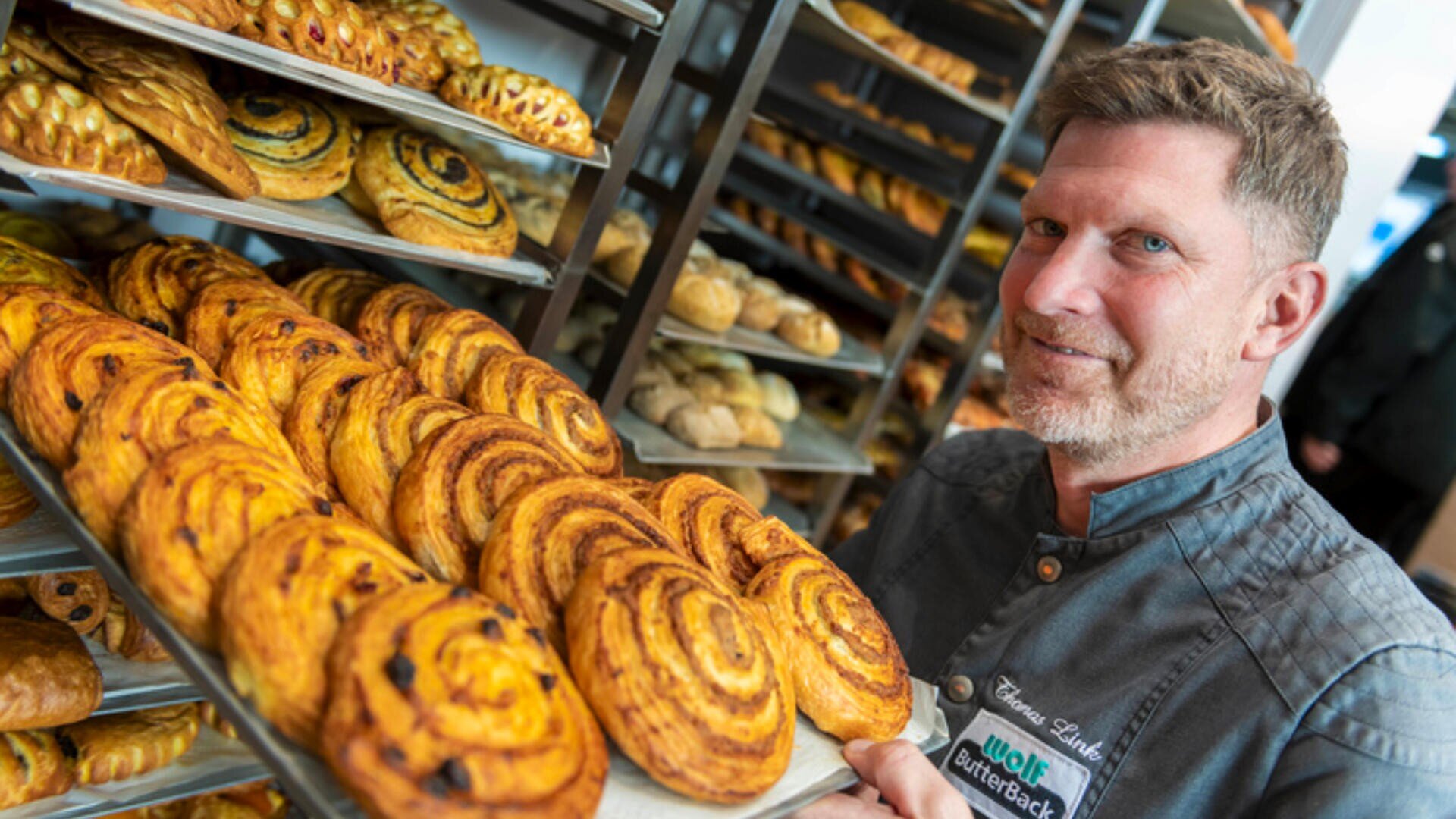 Ein Bäcker in einer grauen Uniform hält ein Tablett mit frisch gebackenem Gebäck mit einem Wirbelmuster und lächelt vor den Regalen mit verschiedenen Backwaren in einer Bäckerei.