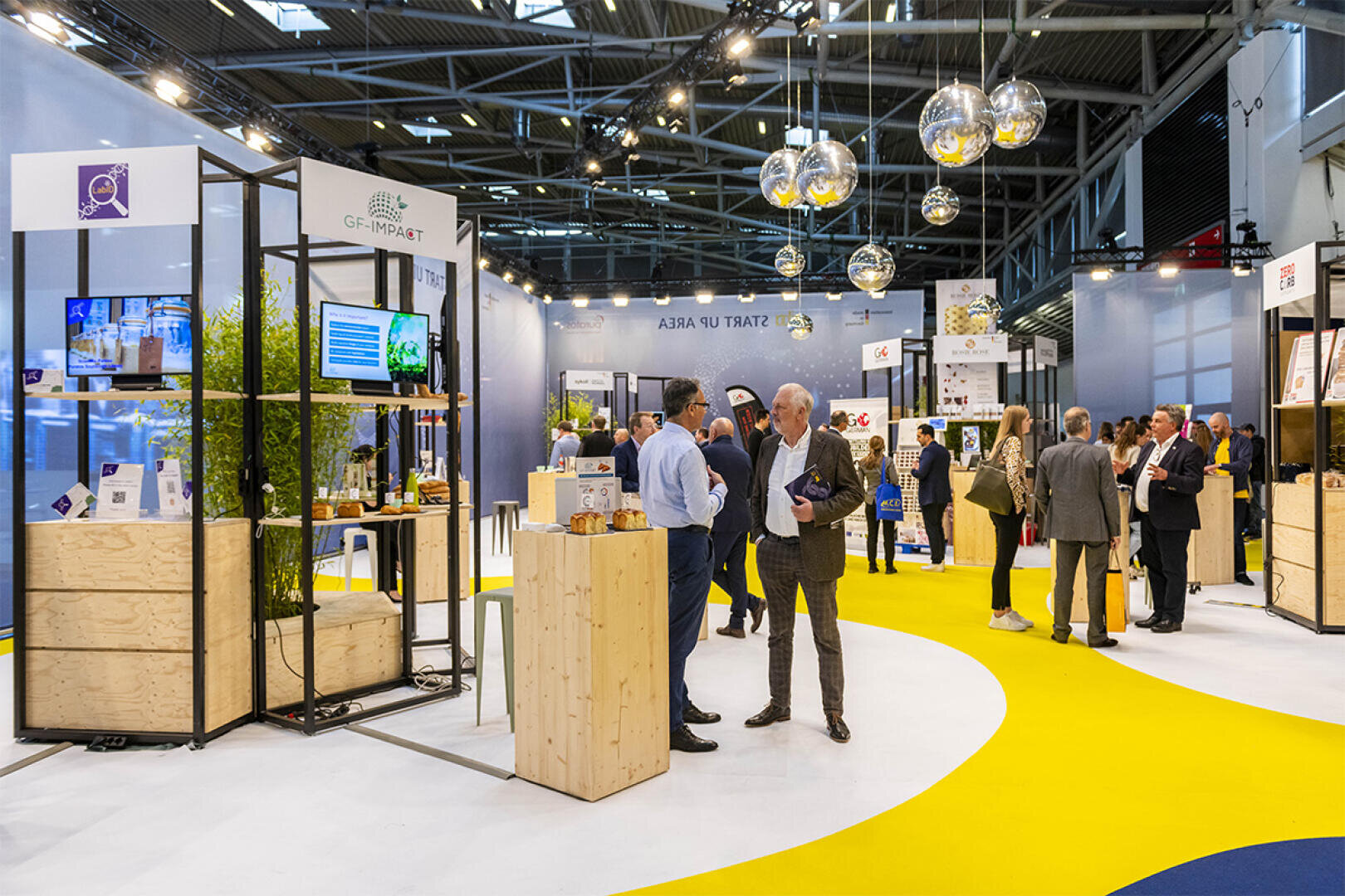 People converse and browse displays at an indoor trade show or exhibition, with modern booths, product samples, and digital screens. The space has bright lighting, yellow and white floor patterns, and hanging spherical lights.