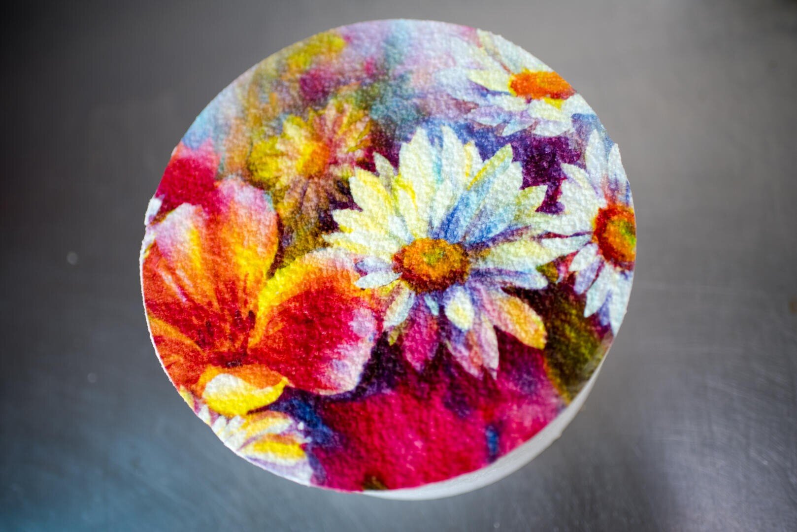 A round cake decorated with a colorful, realistic floral design featuring white daisies and vibrant red, pink, and yellow flowers against a blurred background.