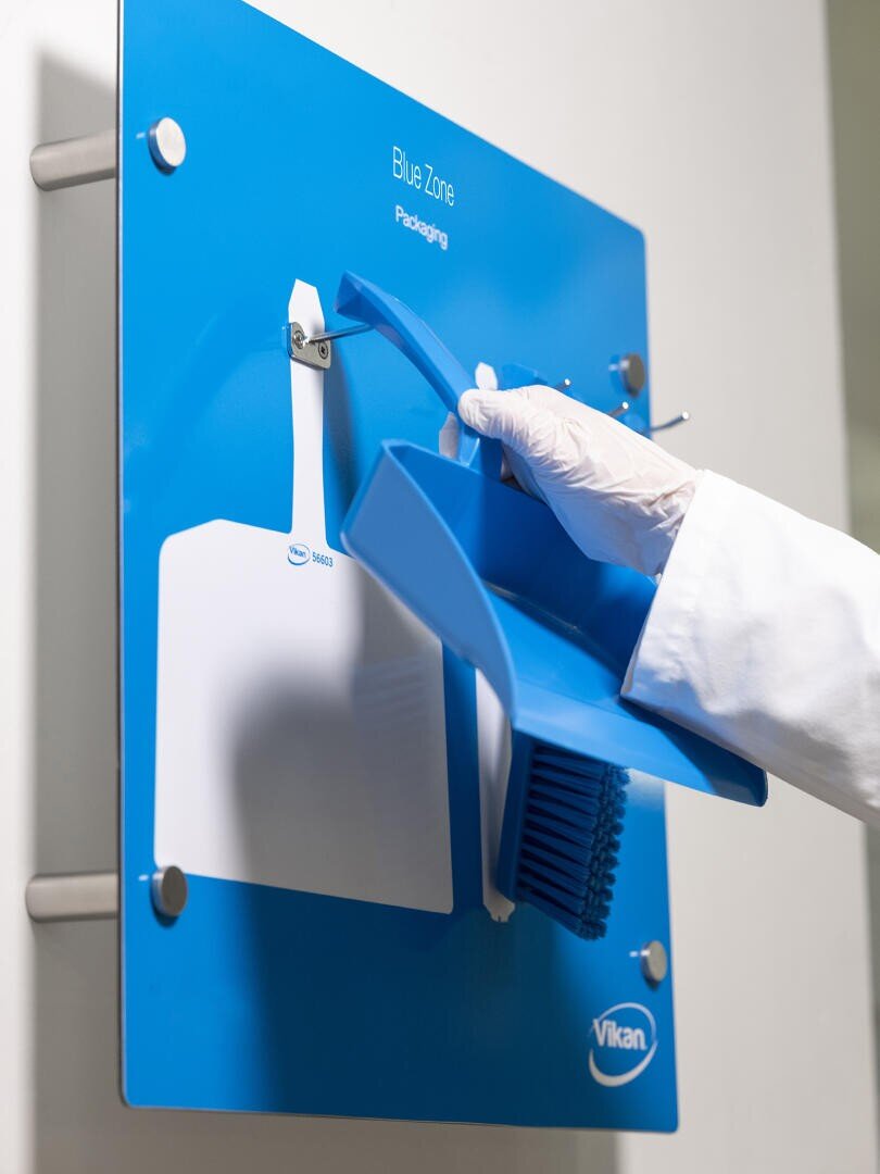 Eine behandschuhte Hand stellt eine blaue Kehrschaufel auf eine blaue, an der Wand befestigte Reinigungsstation mit der Aufschrift Blue Zone Packaging, neben einer weißen Kehrschaufel und einem blauen Besen.