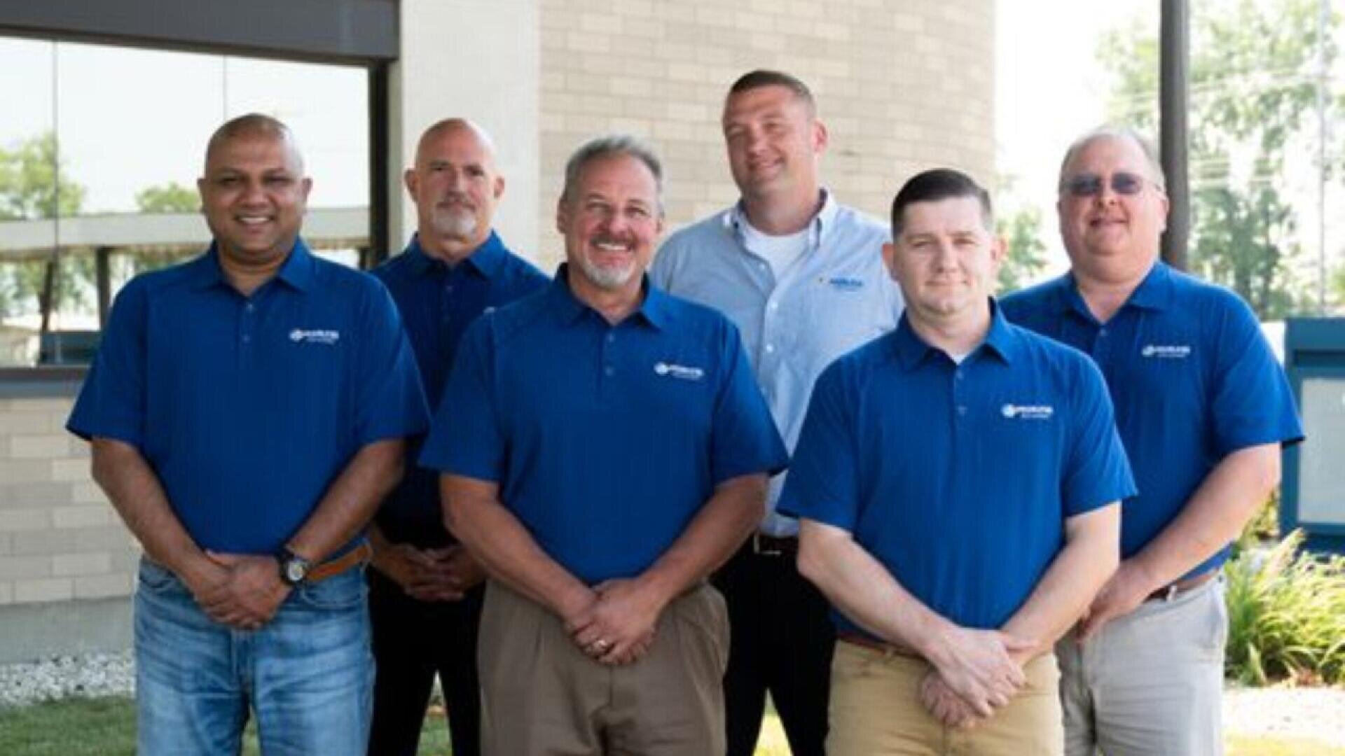 Sechs Männer stehen im Freien vor einem Gebäude, alle tragen passende blaue Poloshirts und lächeln in die Kamera. Im Hintergrund sind Fenster, Ziegelsteine und Grünflächen zu sehen.