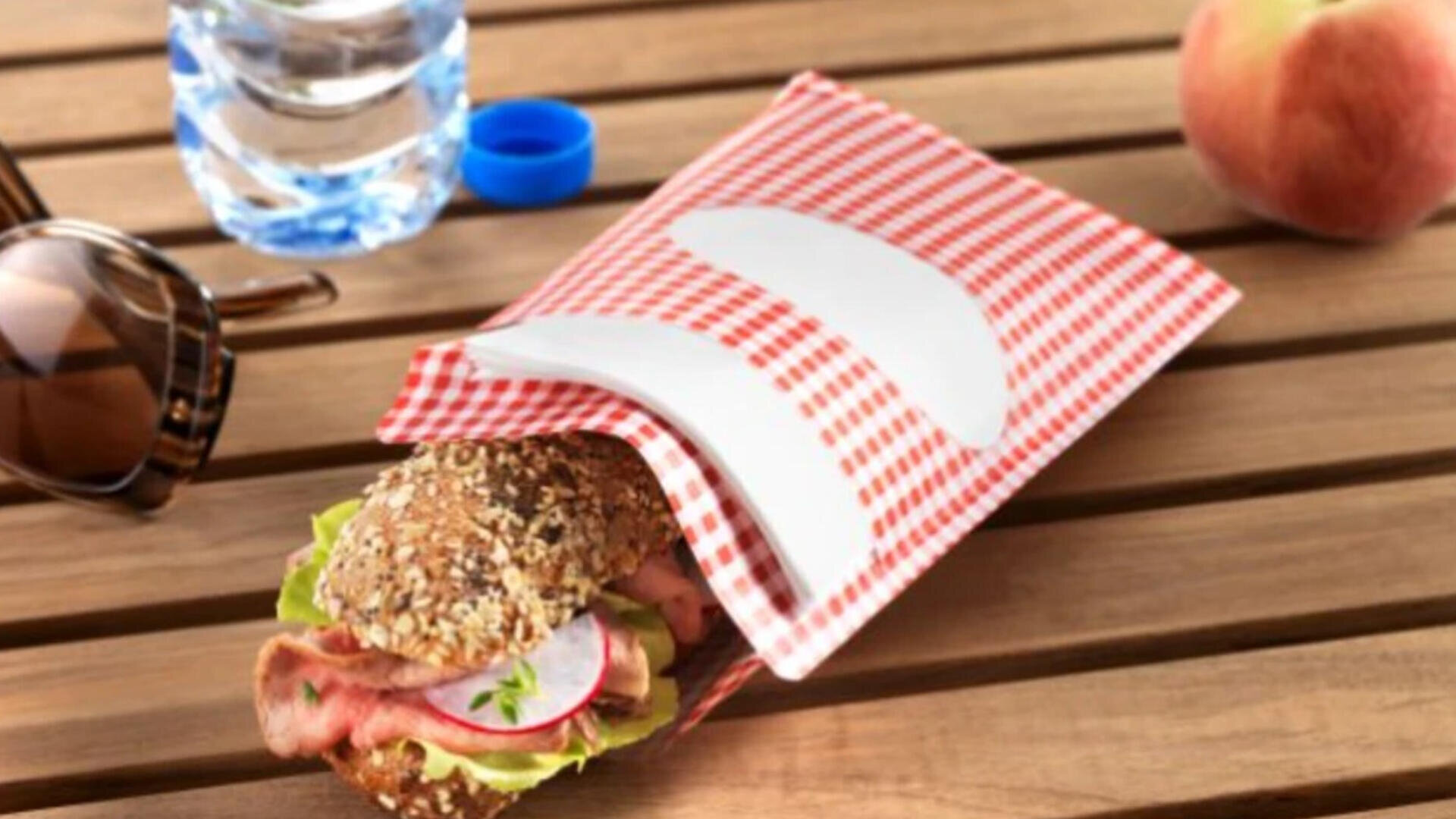 A multigrain sandwich with lettuce, radish, and meat in a red-and-white checkered paper bag on a wooden table, next to sunglasses, a bottle of water, and a peach.