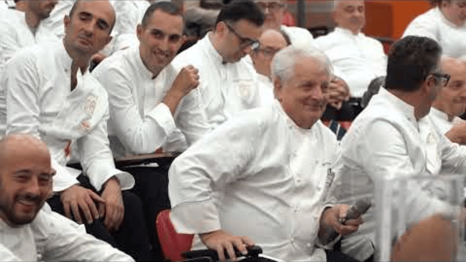 A group of chefs in white uniforms sit closely together in rows of red chairs, smiling and interacting with each other in what appears to be a conference or seminar setting.