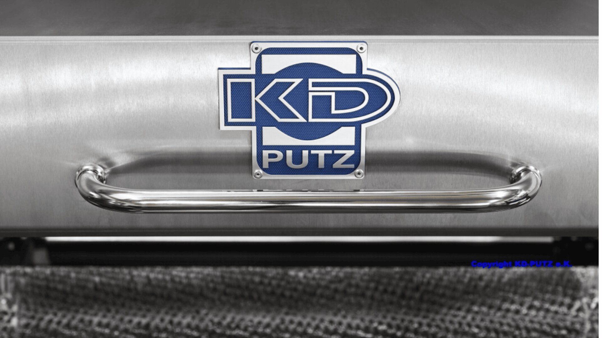 Close-up of a metallic surface with a blue and white KD PUTZ logo above a shiny metal handle. The background shows a textured, mesh-like material and a small copyright notice in blue text.