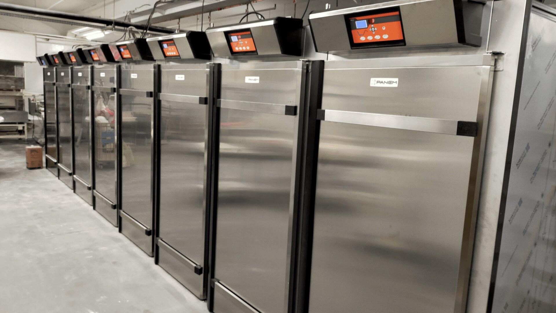A row of large, stainless steel commercial refrigerators or freezers with digital control panels lined up against a wall in an industrial kitchen or storage area.