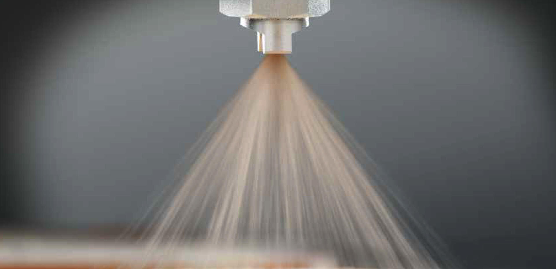 Nahaufnahme einer Sprühdüse, die vor einem neutralen Hintergrund einen feinen Flüssigkeitsnebel oder -strahl abgibt. Der Sprühnebel verteilt sich gleichmäßig kegelförmig.