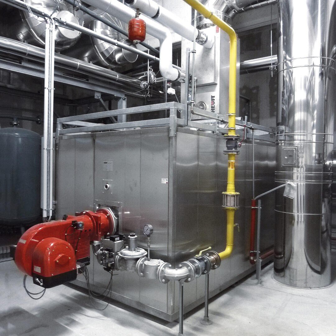 Ein großes industrielles Kesselsystem mit Edelstahlverkleidungen, sichtbaren Rohren, einer roten Brennereinheit, einer gelben Gasleitung und verschiedenen Ventilen in einer sauberen Fabrik oder einem Versorgungsraum mit Metalltanks und -kanälen im Hintergrund.
