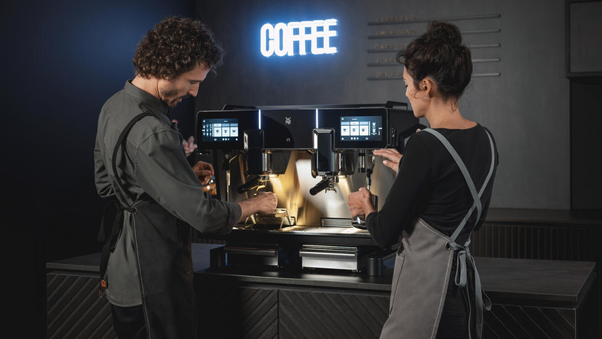 Zwei Baristas in Schürzen bedienen eine große, moderne Kaffeemaschine in einem schwach beleuchteten Café, an der Wand hinter ihnen leuchtet ein KAFFEE-Schild.