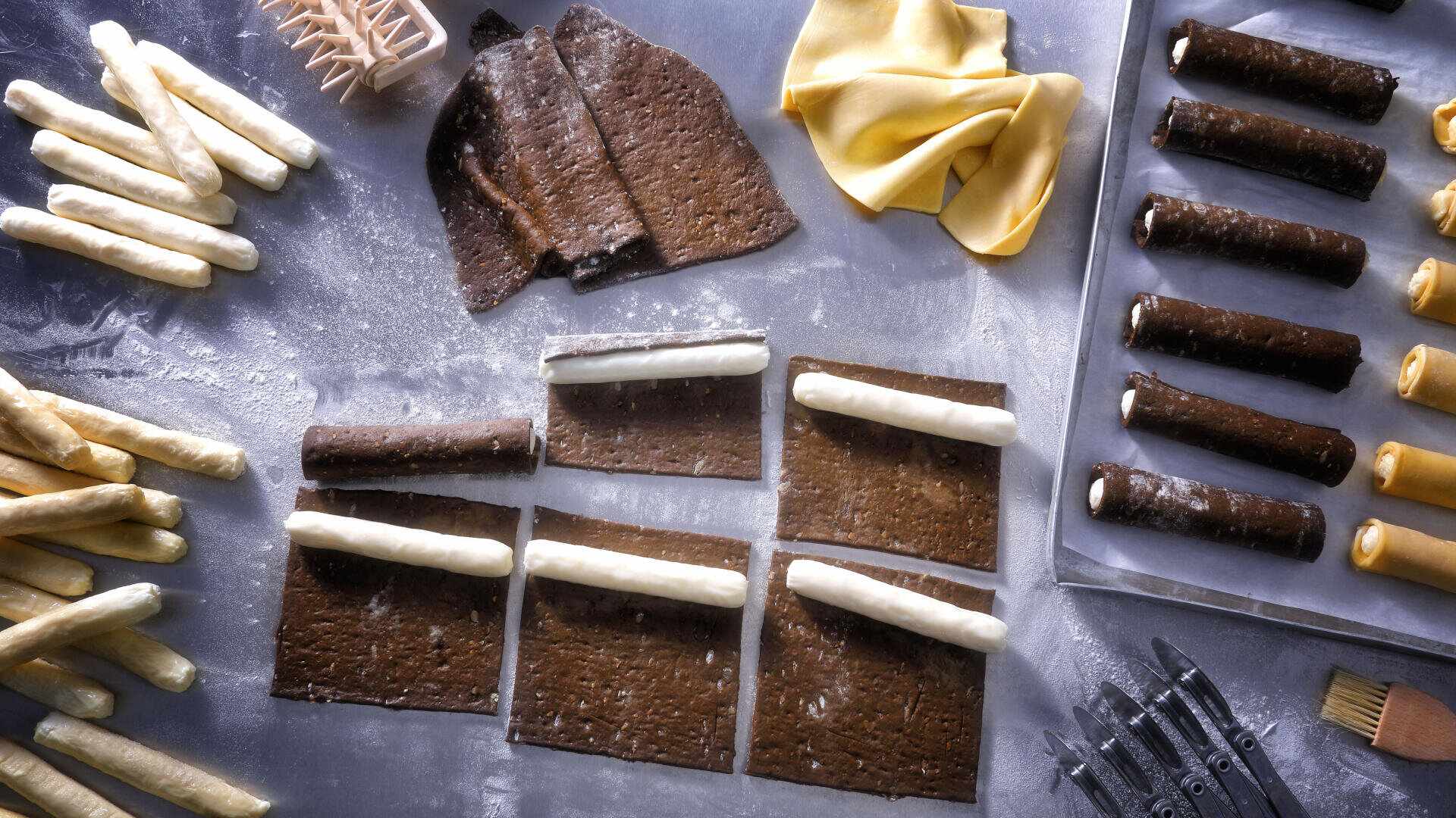 Various dough sheets and strips are laid out on a floured surface, with rolled pastry sticks being wrapped in chocolate and plain dough, and baking tools are scattered around for preparation.