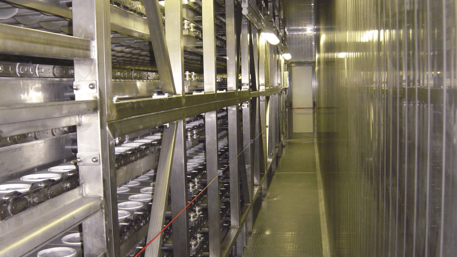 A long, narrow corridor in an industrial facility with metal shelves holding rows of cylindrical containers, possibly part of a storage or manufacturing system, and bright overhead lighting.