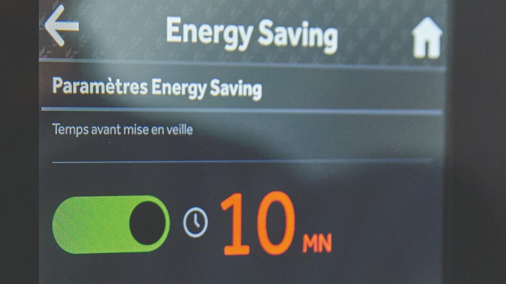 Ein digitaler Bildschirm zeigt die Energiespareinstellungen in französischer Sprache an, mit einem grünen Kippschalter und einem auf 10 Minuten eingestellten Timer, bevor der Schlafmodus aktiviert wird. In der oberen rechten Ecke befindet sich ein Haussymbol.