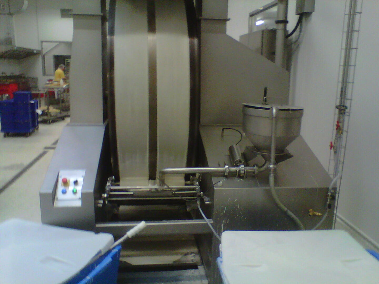 Eine große industrielle Lebensmittelverarbeitungsmaschine mit einem Förderband, einem Bedienfeld und Metallkomponenten in einer sauberen Fabrikumgebung. Blaue Lagerkästen und Geräte aus Edelstahl sind zu sehen.