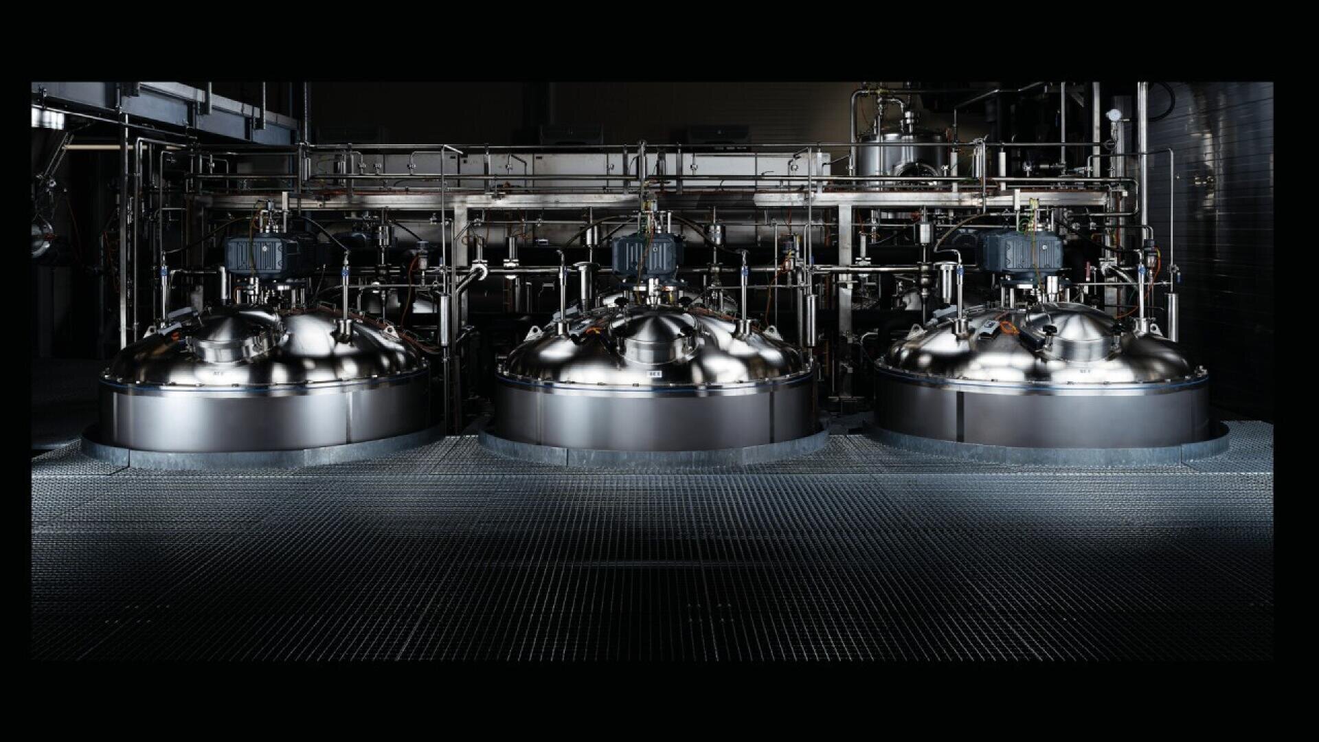 Three large, shiny, stainless steel industrial tanks with pipes, gauges, and machinery mounted above them in a dark, modern factory setting.
