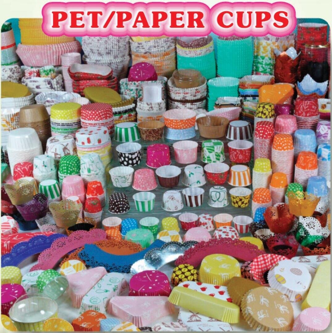 A large selection of colorful PET and paper baking cups in various sizes, patterns, and shapes are presented in rows on a table under a pink banner reading “PET/PAPER CUPS.”.