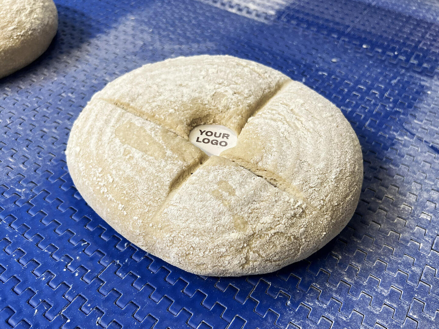 Ein runder, bemehlter Teiglaib mit einem Kreuzschnitt auf einer blauen, strukturierten Oberfläche. In der Mitte des Teigs befindet sich ein kleines Stück Papier mit der Aufschrift „IHRES LOGO“.