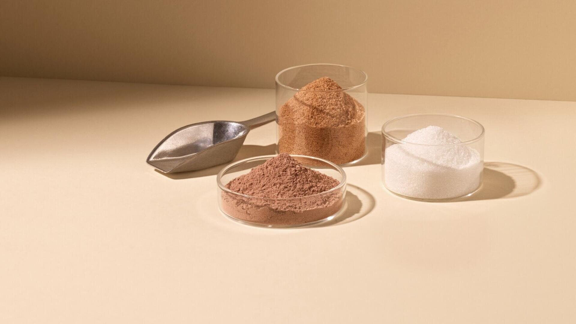 Three glass containers hold different powders: a tall glass with brown sugar, a small dish with cocoa powder, and a small glass bowl with white sugar. A metal scoop rests next to them on a beige background.