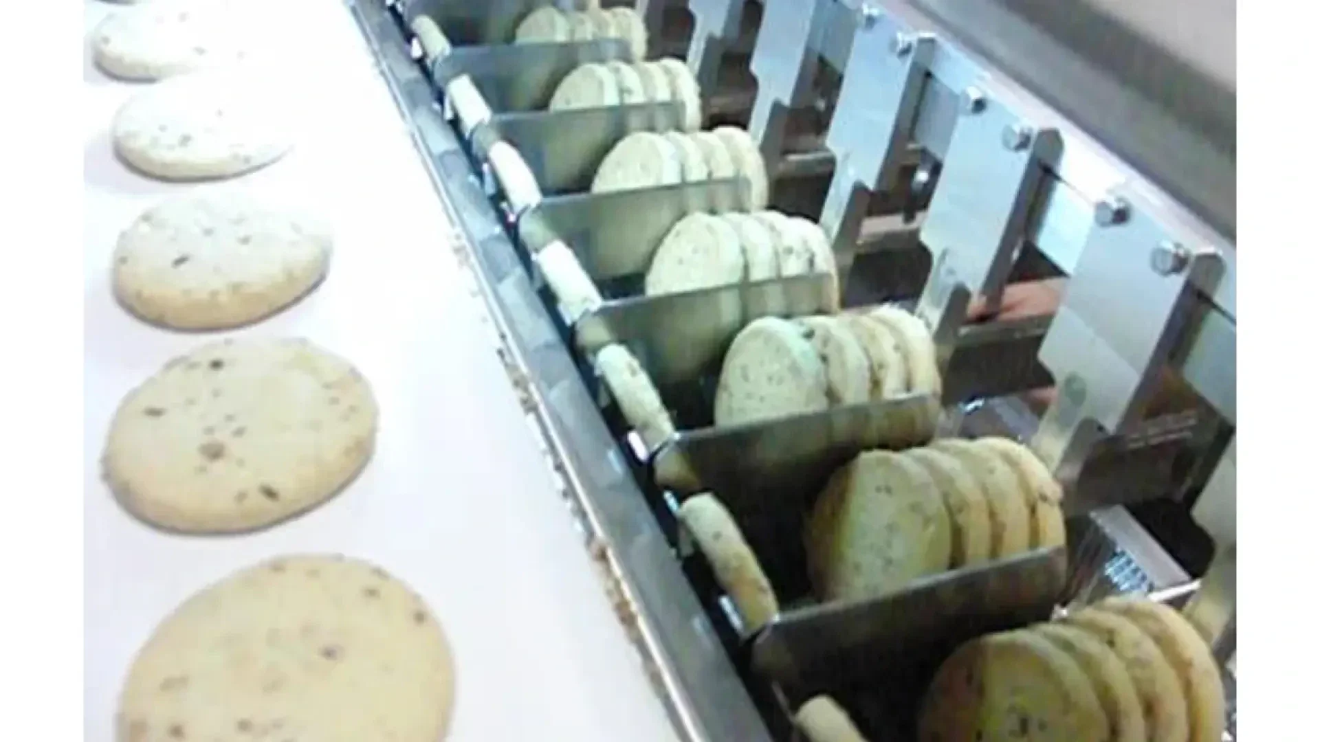 A cookie production machine sorting and aligning rows of round cookies on a conveyor belt, with some cookies already placed on a white surface nearby.