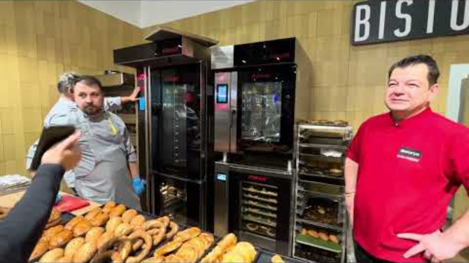 Two men stand behind a counter filled with various breads and pastries. One man wears a gray chefs coat, the other a red shirt. Behind them are ovens and racks. A person in the foreground holds up a phone to take a photo.