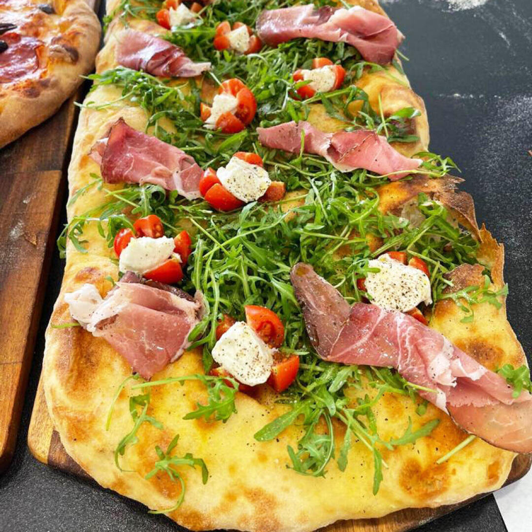 A rectangular flatbread topped with fresh arugula, cherry tomato halves, slices of prosciutto, and dollops of creamy cheese, with a sprinkle of black pepper on a wooden board.
