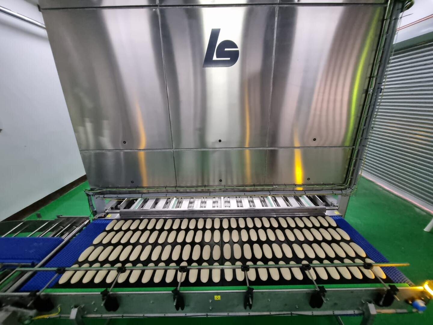 Eine Fabrikmaschine produziert Reihen von gleichmäßig geformten ovalen Teiglingen auf einem blauen Förderband, über dem eine große Metalltafel mit den Buchstaben L und S angebracht ist.